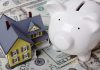 A model house next to a white piggy bank on a bed of dollar bills