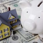 A model house next to a white piggy bank on a bed of dollar bills