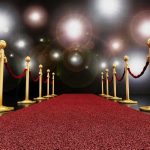 A red carpet lined with gold stanchions and ropes under bright spotlights