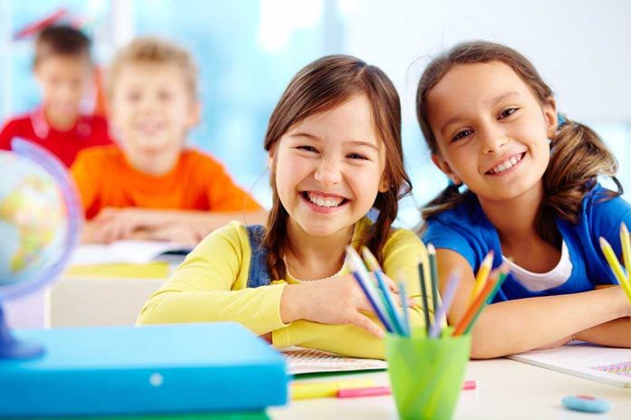 Two smiling girls in a classroom with colorful stationery and classmates in the background