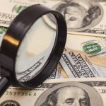 A magnifying glass resting on a pile of U.S. dollar bills
