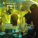 armed officer confronts people in hazmat suits inside a lab