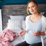 Pregnant woman sitting on bed holding pills and a glass of water