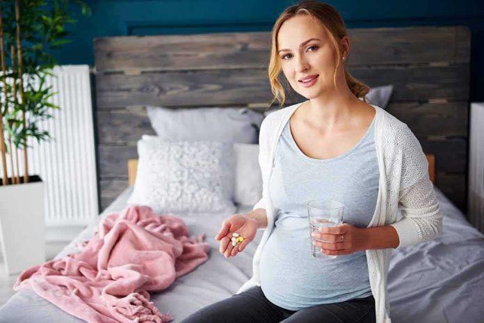 shutterstock_1100580743 Pregnant woman sitting on bed holding pills and a glass of water