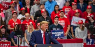 Donald Trump speaks at a rally podium; Christian Jones and Bear Pascoe are in the crowd behind him